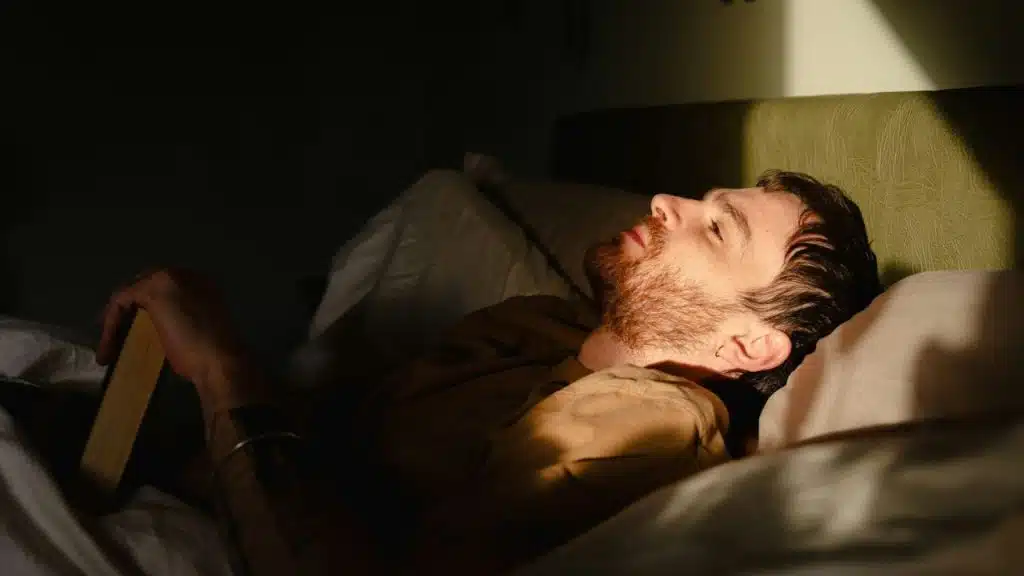 A bearded man lies in bed, looking up with his eyes closed, partially lit by a light.