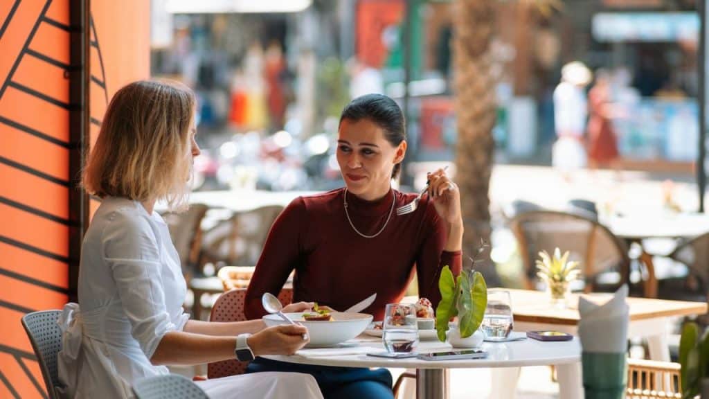 Two women are seated at an outdoor restaurant table, eating and talking.