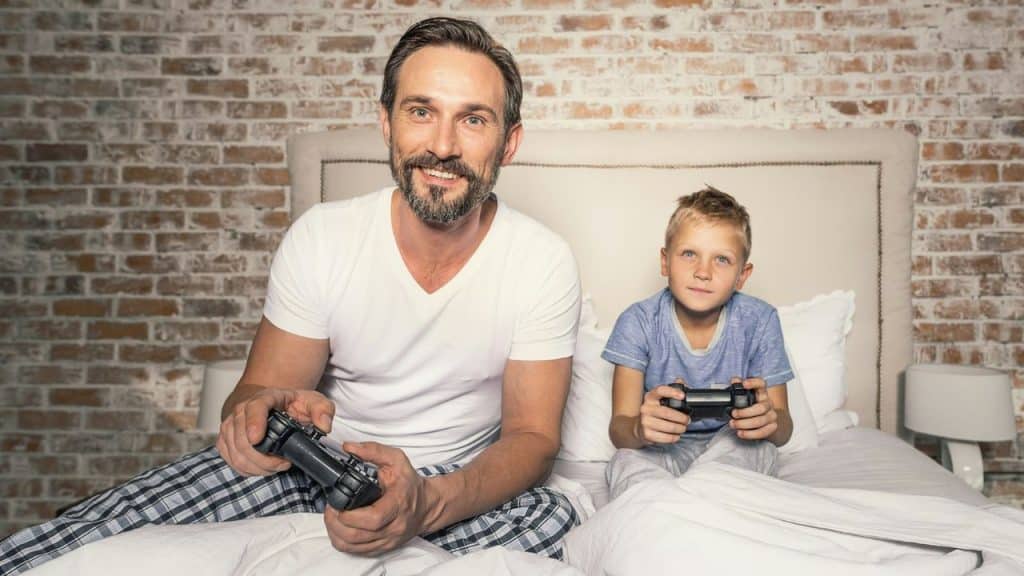 A smiling man and a young boy sit on a bed, holding video game controllers in front of a brick wall.