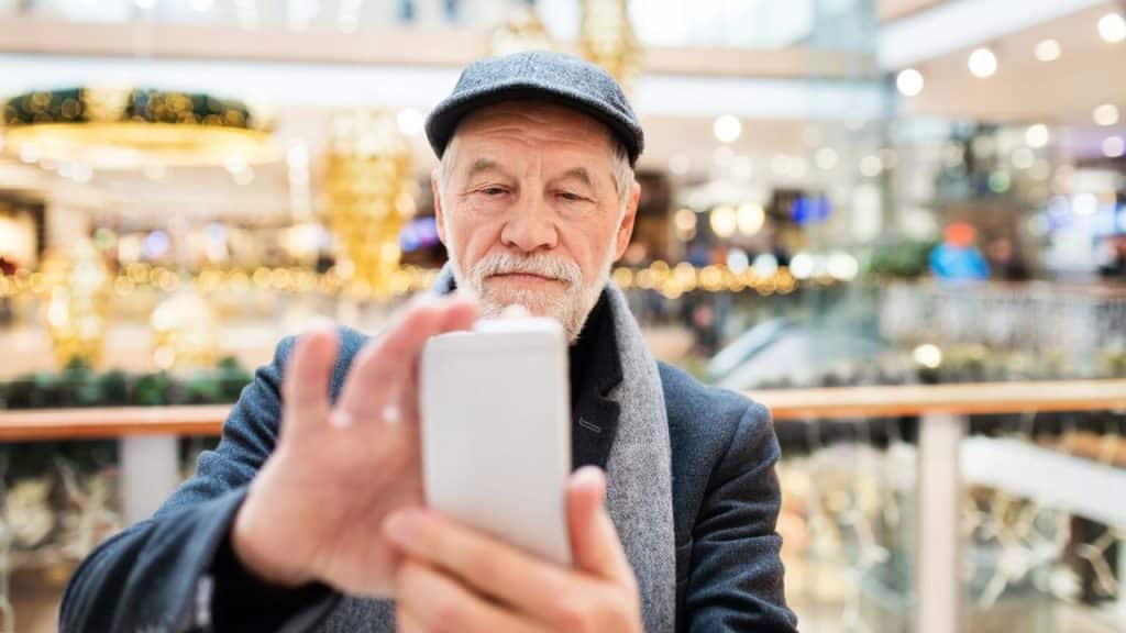 Older man in a cap and scarf looking at his smartphone in a busy mall.