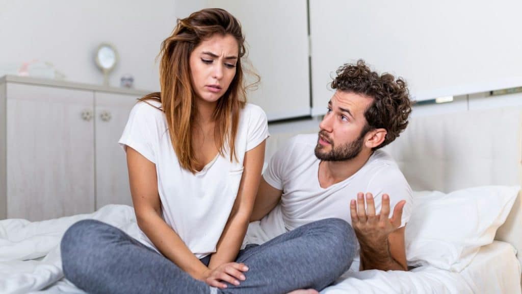 A woman looking unhappy on a bed while a man gestures at her.