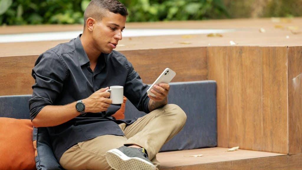 A man sitting outdoors holding a coffee cup and looking at his smartphone.
