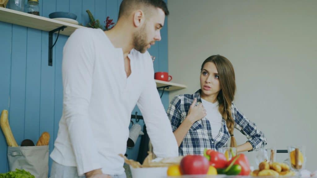 A man in a white shirt leans over a counter facing a woman in a plaid shirt who looks upset and is gesturing in a kitchen.