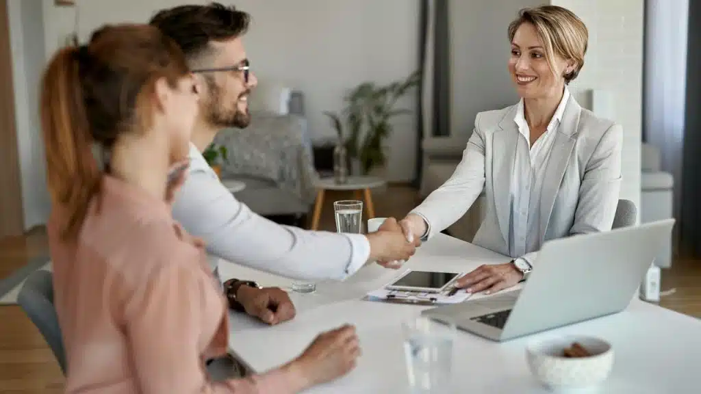 A smiling businesswoman shaking hands with a man while sitting with a couple at a desk.