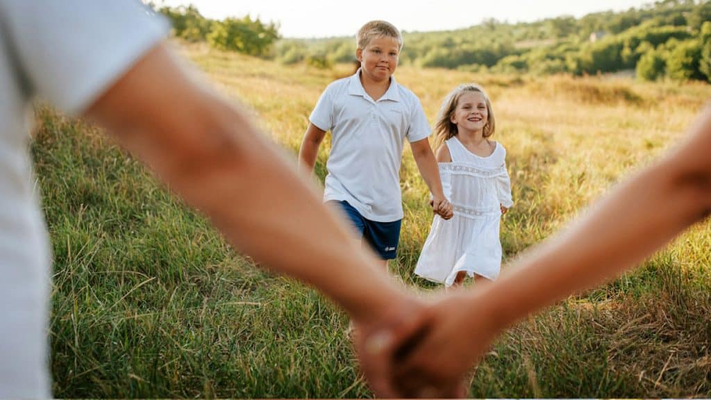 Two kids walking in the field while their parents are holding each other's hands.