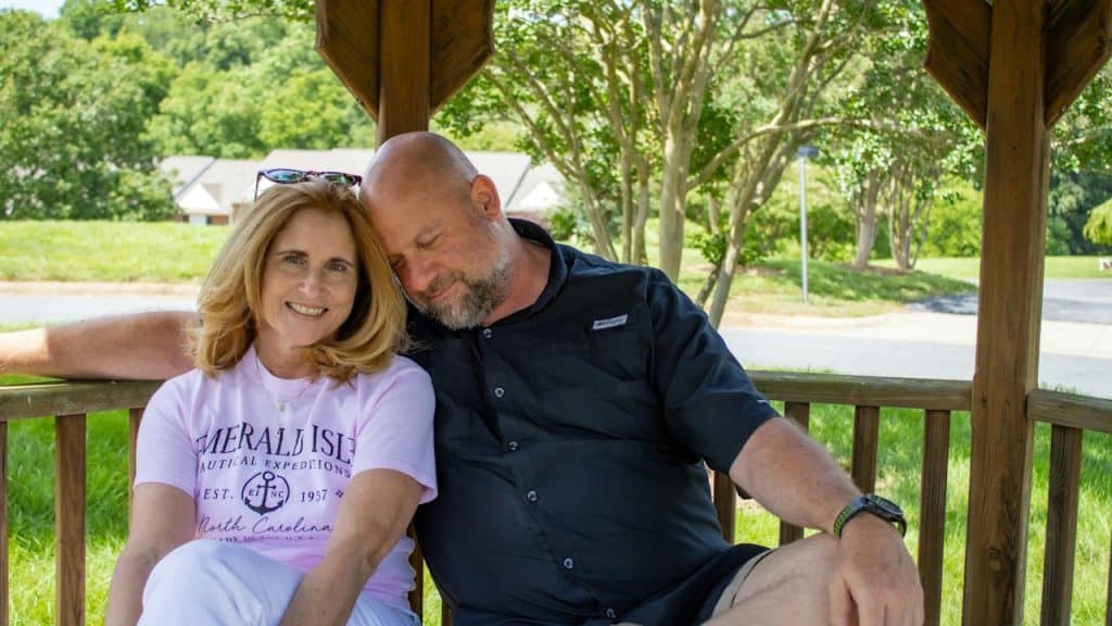A happy middle-aged couple sitting close together in a wooden gazebo, with the man resting his head on the woman's shoulder.
