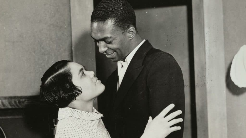 A black and white photo of a Black man in a suit smiling down at a woman with short hair who is looking up at him.