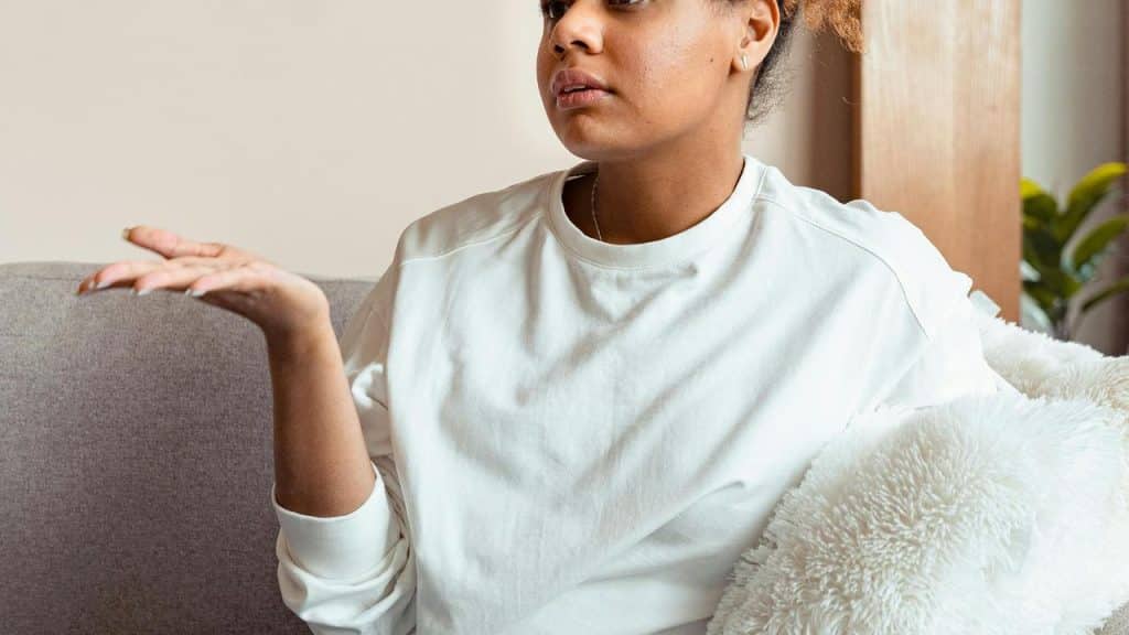 A woman sitting on a couch with her hand raised in a questioning gesture.