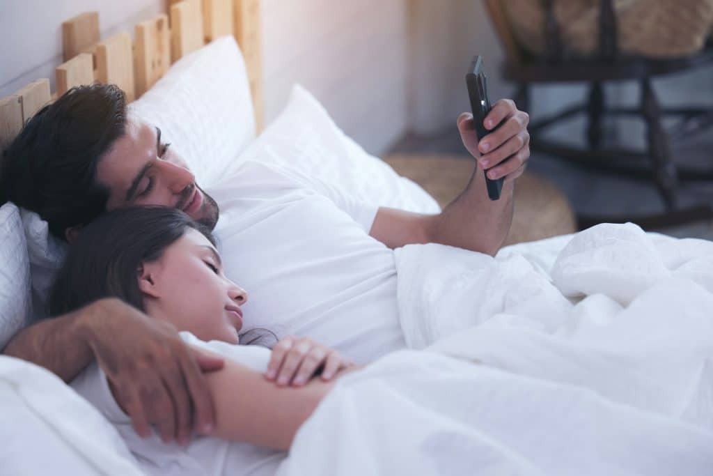 The man is using his mobile phone while the woman is sleeping beside him.