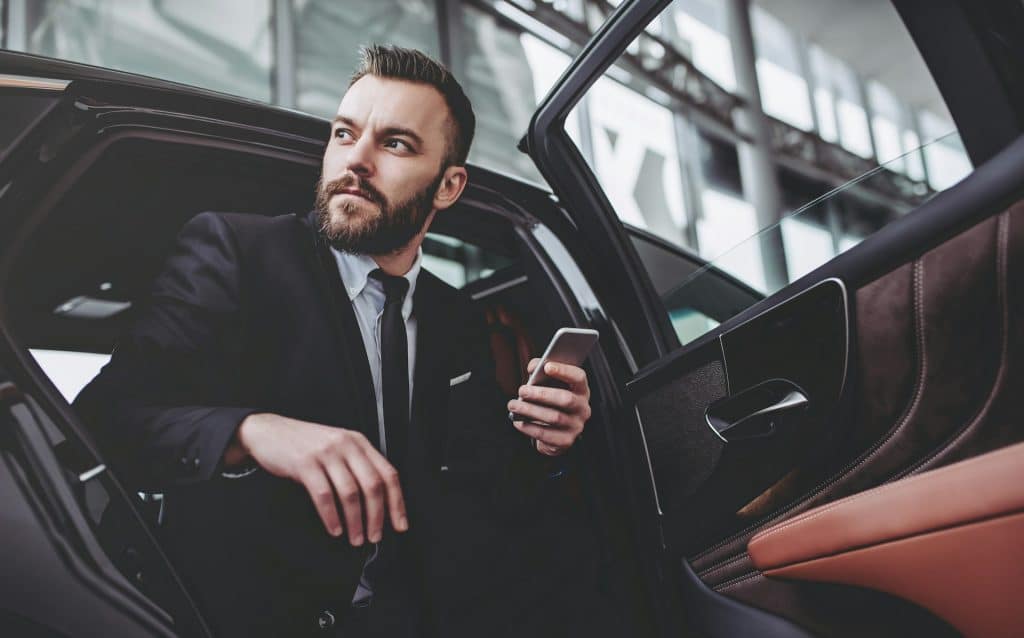 The businessman is sitting in his car and holding a smartphone.