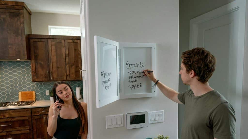 A man and a woman standing in the kitchen and writing about things to purchase.