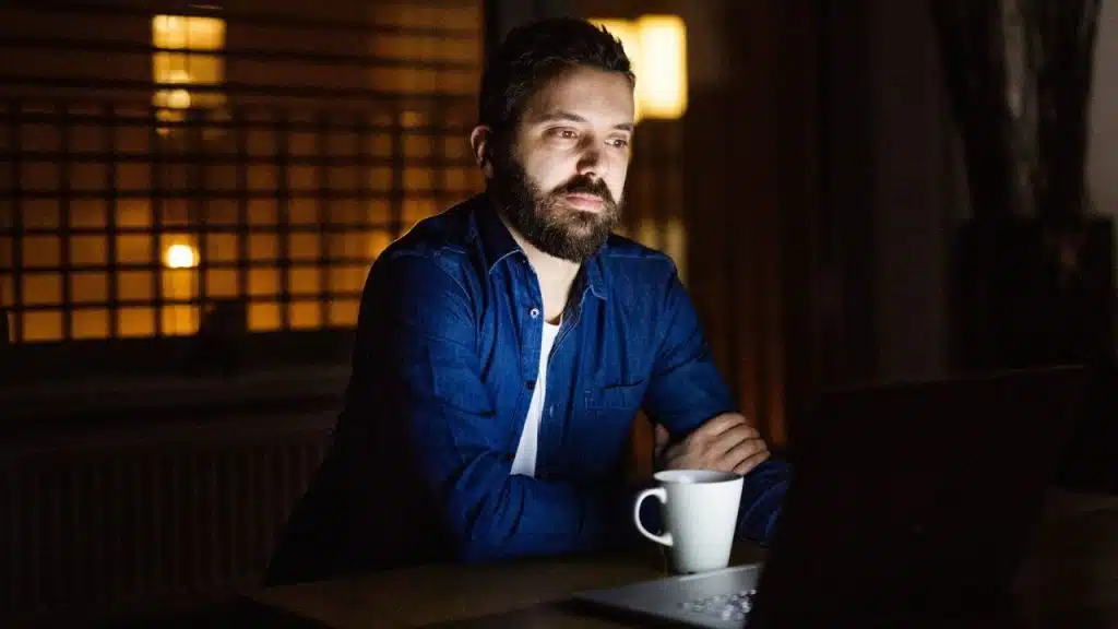 A bearded man in a blue denim shirt sits at a desk in the dark, looking at a laptop screen. A white mug is in front of him.