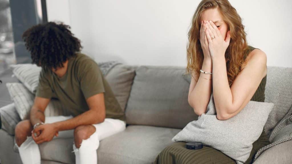A woman covering her face on a couch while a man sits beside her facing away.