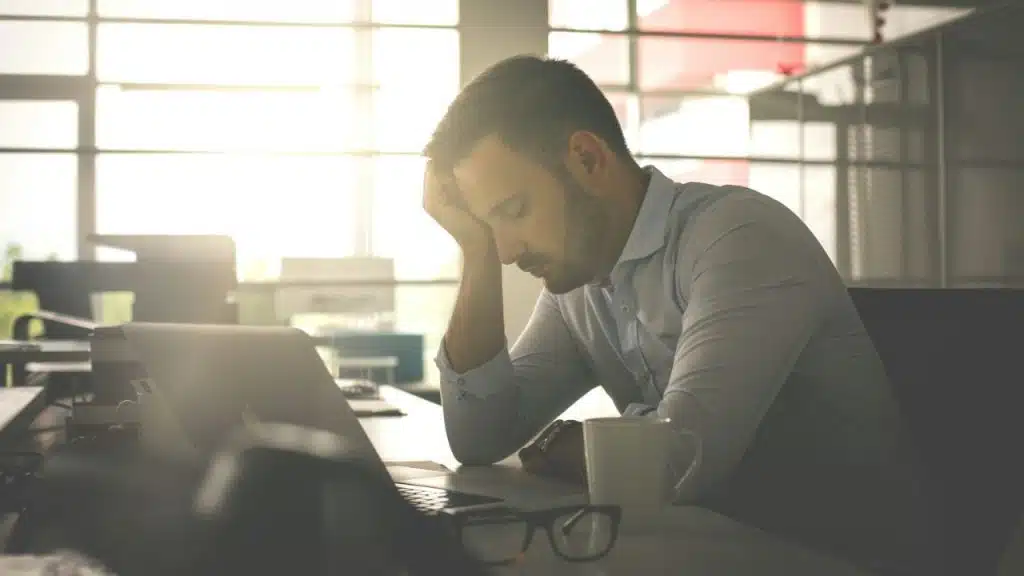 A man is sitting at a desk with his head resting on his hand in a bright office.