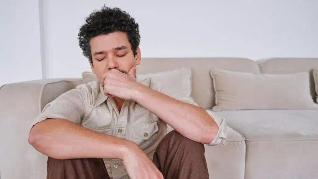 A man with curly hair is sitting on the floor with his eyes closed and hand on his face.
