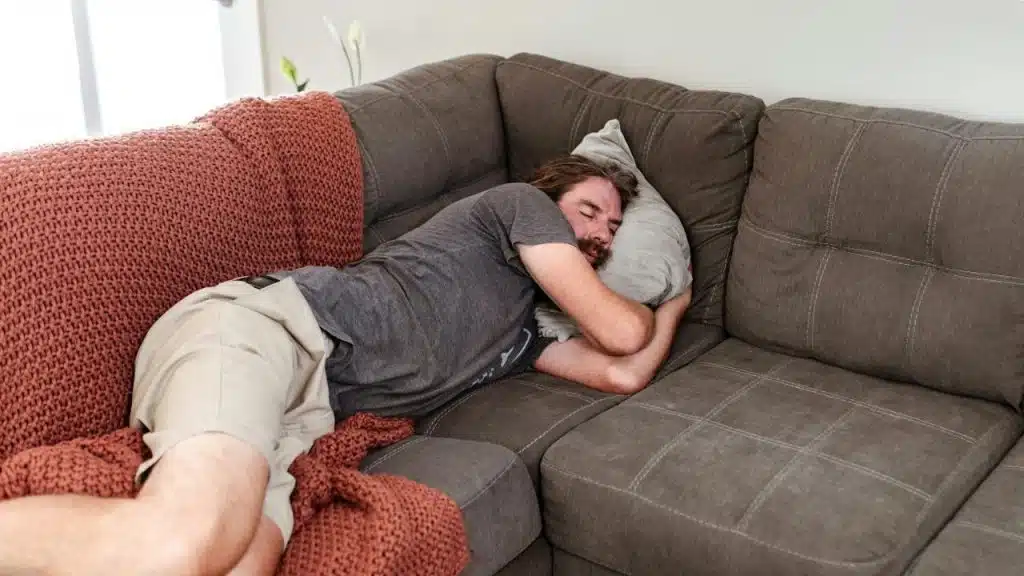 A man is curled up and sleeping on a gray sectional sofa with a reddish-brown blanket.