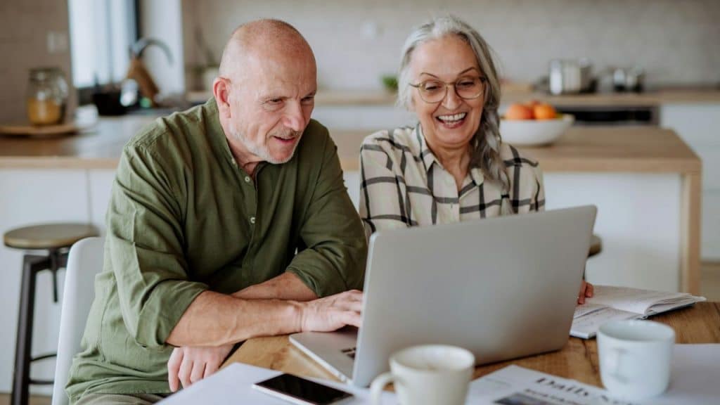 Happy older couple sitting at a kitchen table looking at a laptop together.