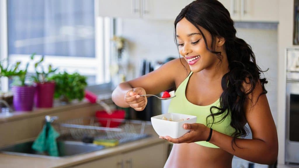 A woman smiling while eating cereal with fruit in a kitchen.