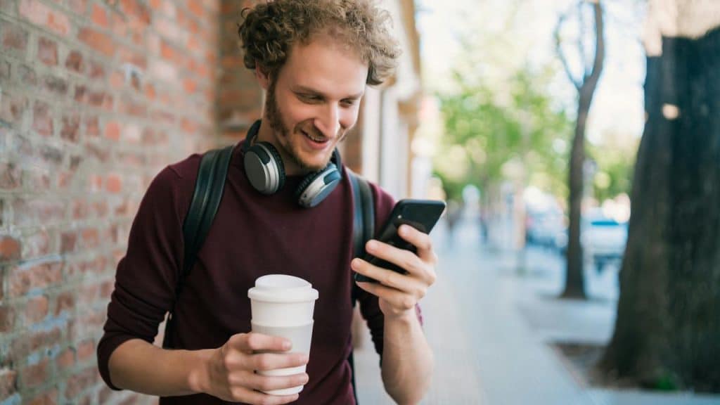 A man smiling at his phone while holding a coffee cup outdoors.