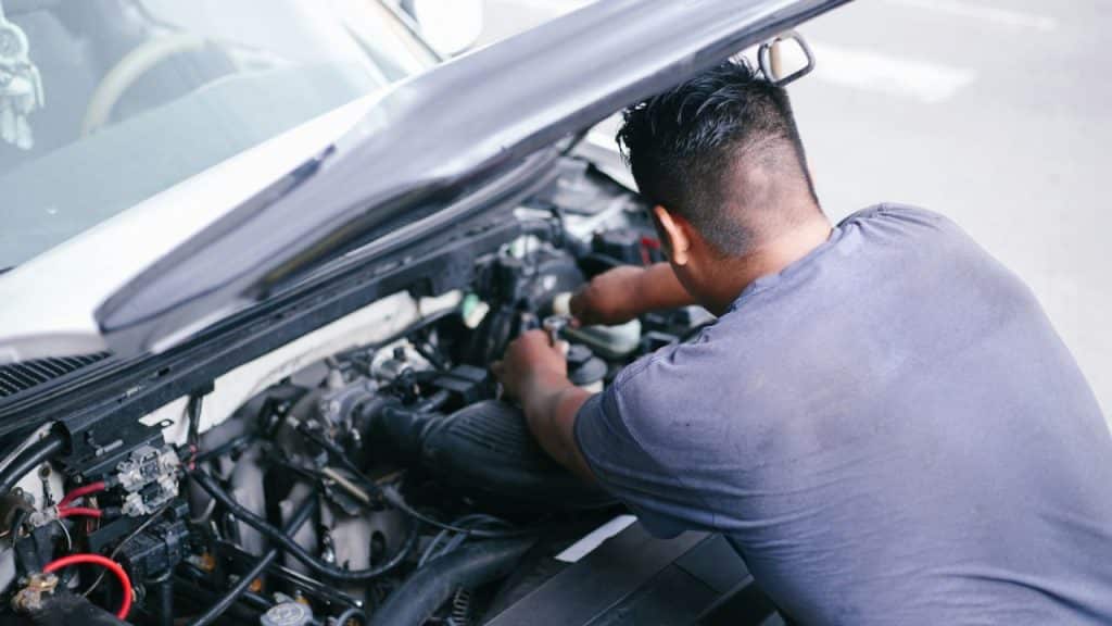 A man in a gray shirt is leaning over an open car hood, working on the engine.