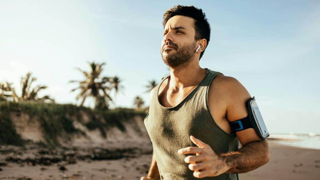 A man wearing earbuds and an armband runs on a sunny beach with palm trees.