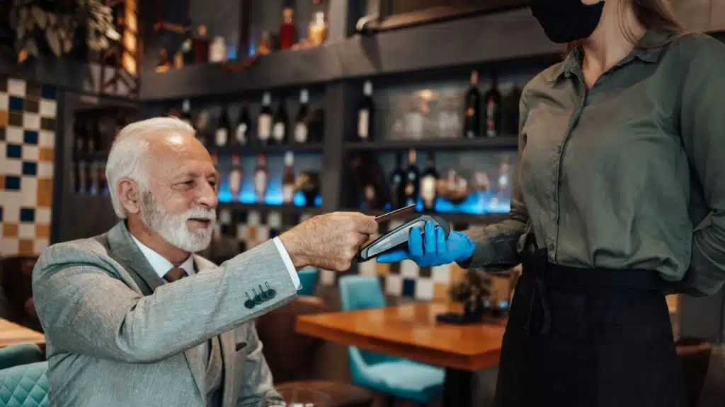 An older man with a white beard is paying with a credit card to a masked waitress.