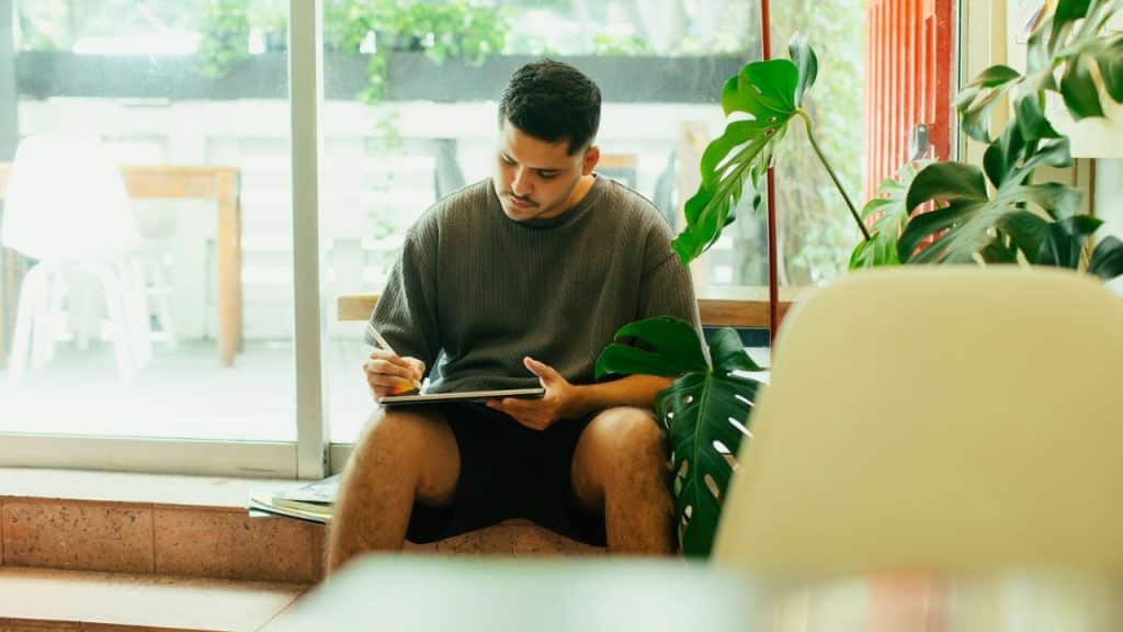 Man sitting by a large window, using a stylus on a tablet.