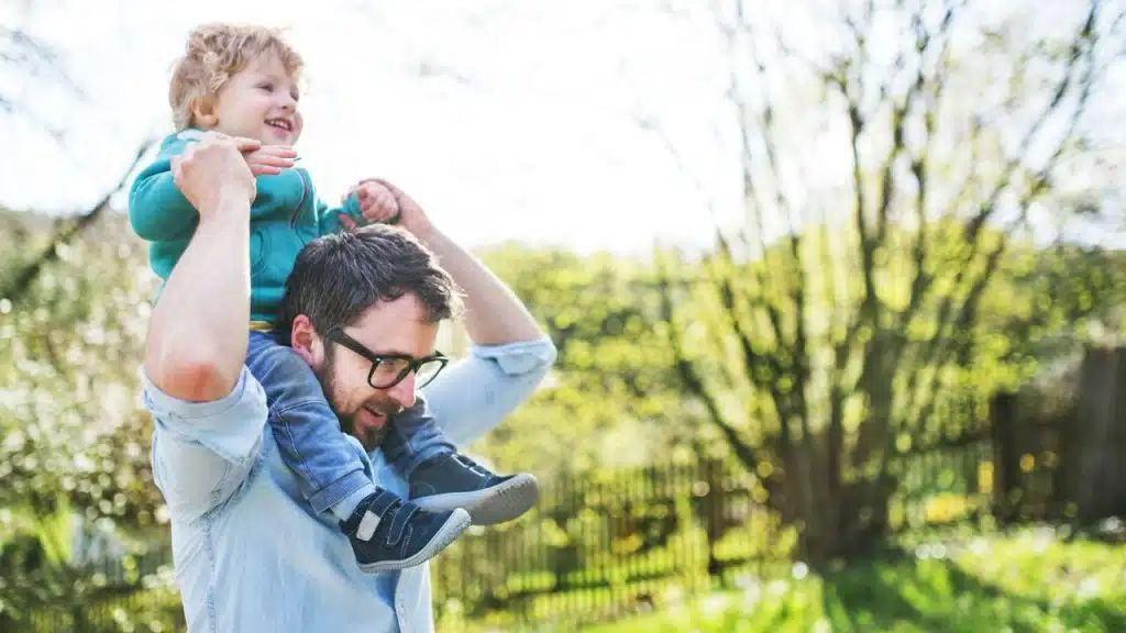 A happy father in glasses is carrying his laughing son on his shoulders outdoors.