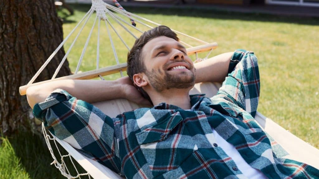 Smiling man relaxing in a hammock outdoors, wearing a green and blue plaid shirt.