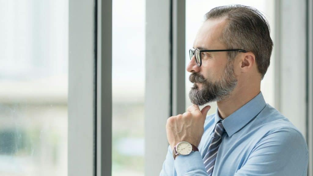 Middle-aged man in a shirt and tie looks thoughtfully out a large window.