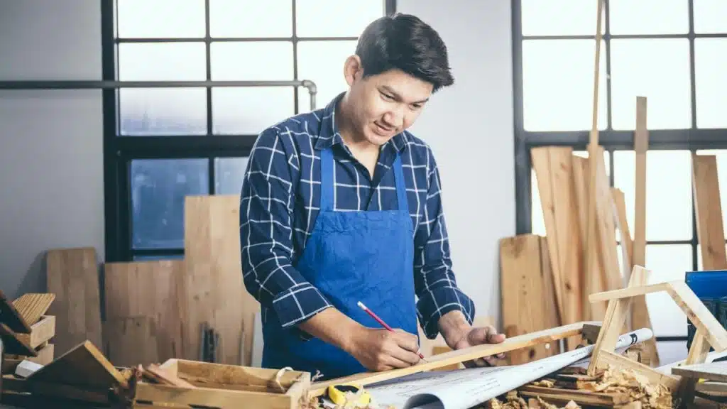 A smiling man in a blue apron is drawing on a piece of wood in a workshop.