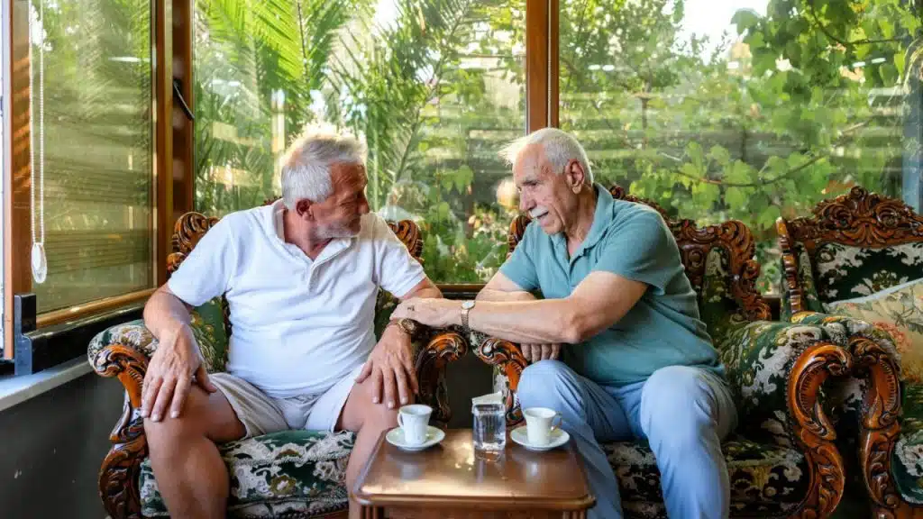 Two older men talk while sitting on ornate sofas next to a large window.