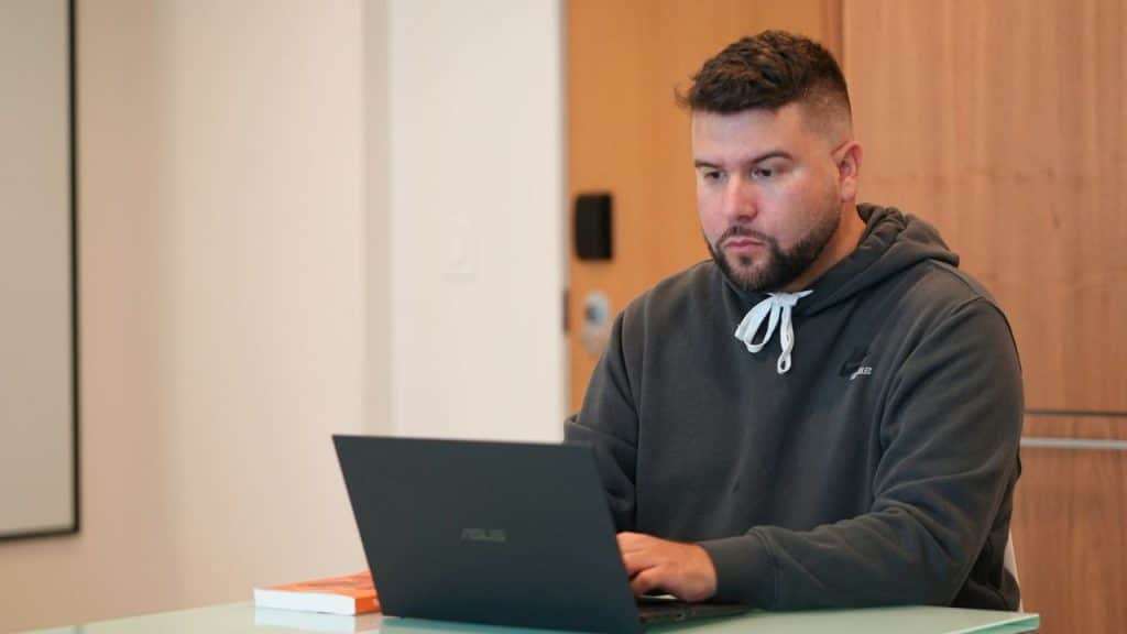 Man in a dark gray hoodie sits at a table and works on a black laptop.