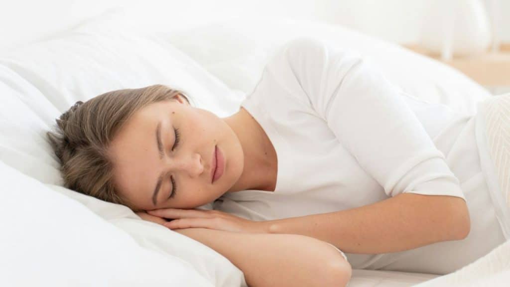 Close-up of a young woman sleeping on her side in white bedding and a white shirt.