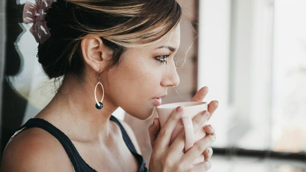 A woman holding a cup close to her face while looking out thoughtfully.