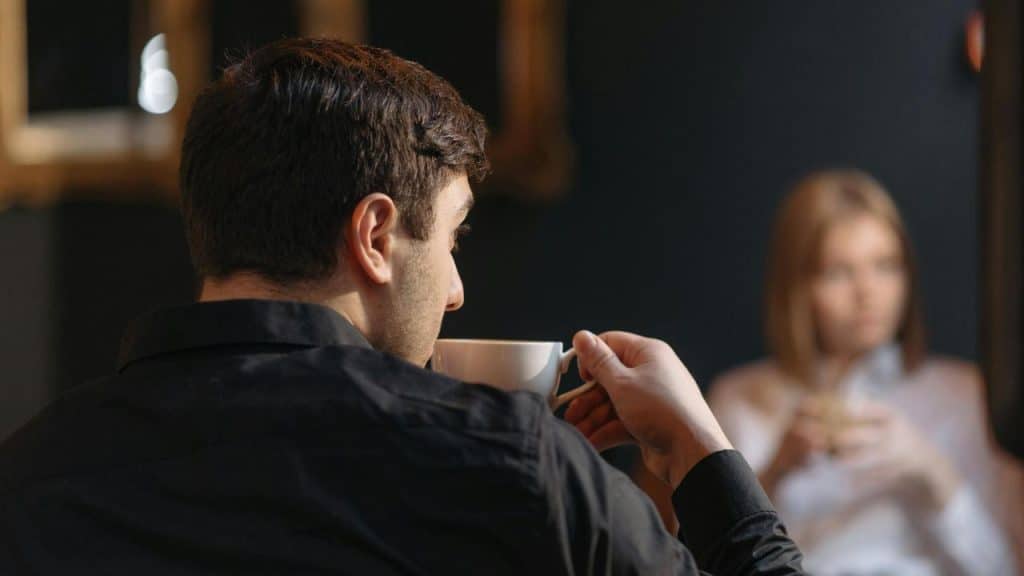 A man in a black shirt drinking coffee with a woman sitting in the background.