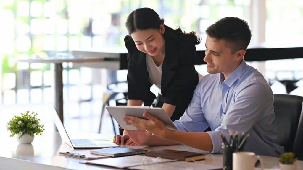 Two office workers are smiling while reviewing a tablet and laptop at a white desk.