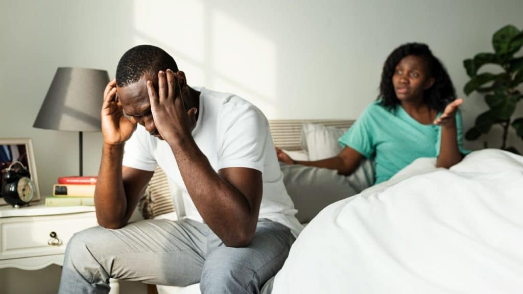 A man sits on a bed with his head in his hands while a woman argues behind him.