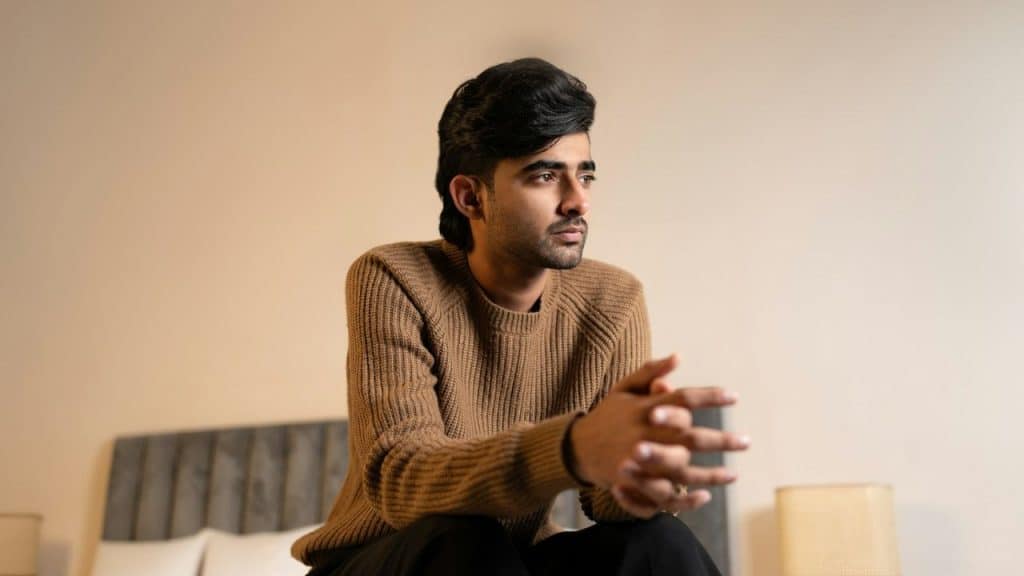 Young man in a brown sweater sitting on a bed, looking off to the side with his hands clasped.