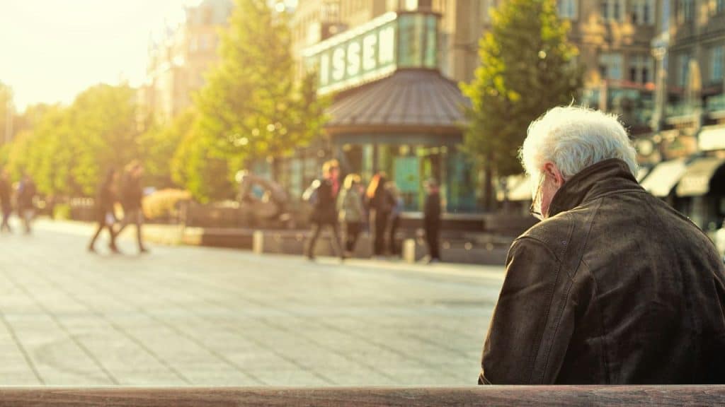 Older person with white hair sitting on a bench in a sunny, busy square.