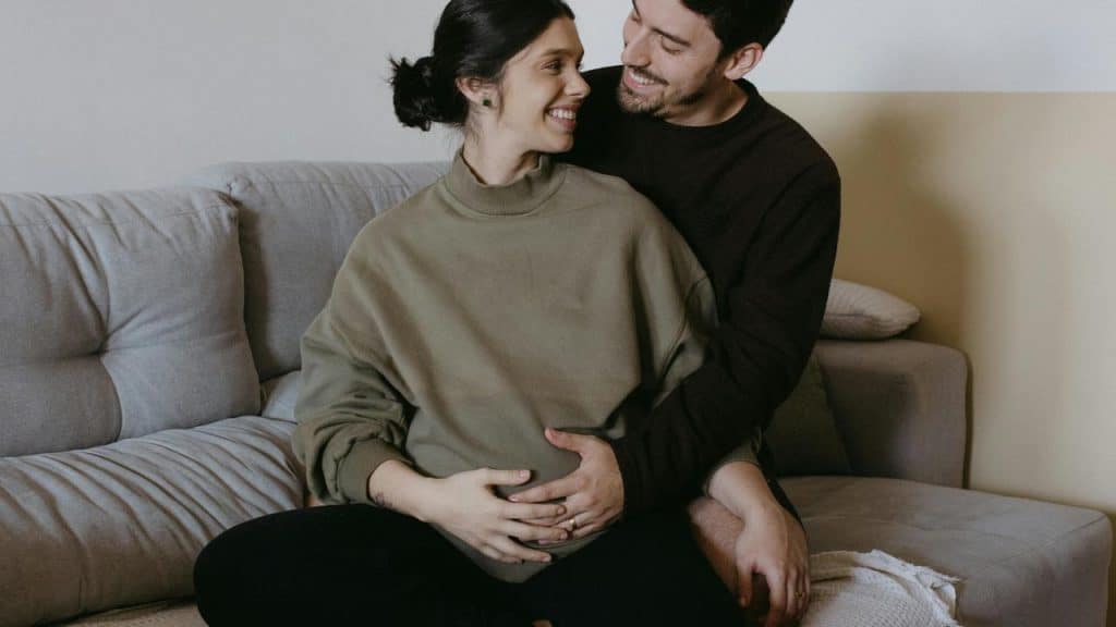 A couple sitting on a couch smiling while the man holds the woman’s pregnant belly.
