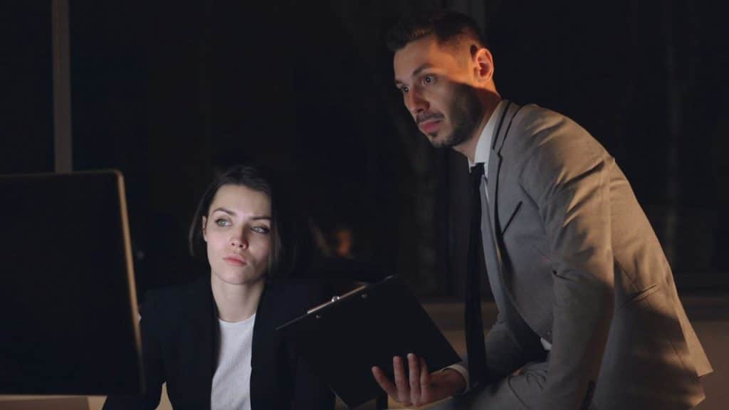 A man in a suit is standing over a woman working on a computer in a dark office.