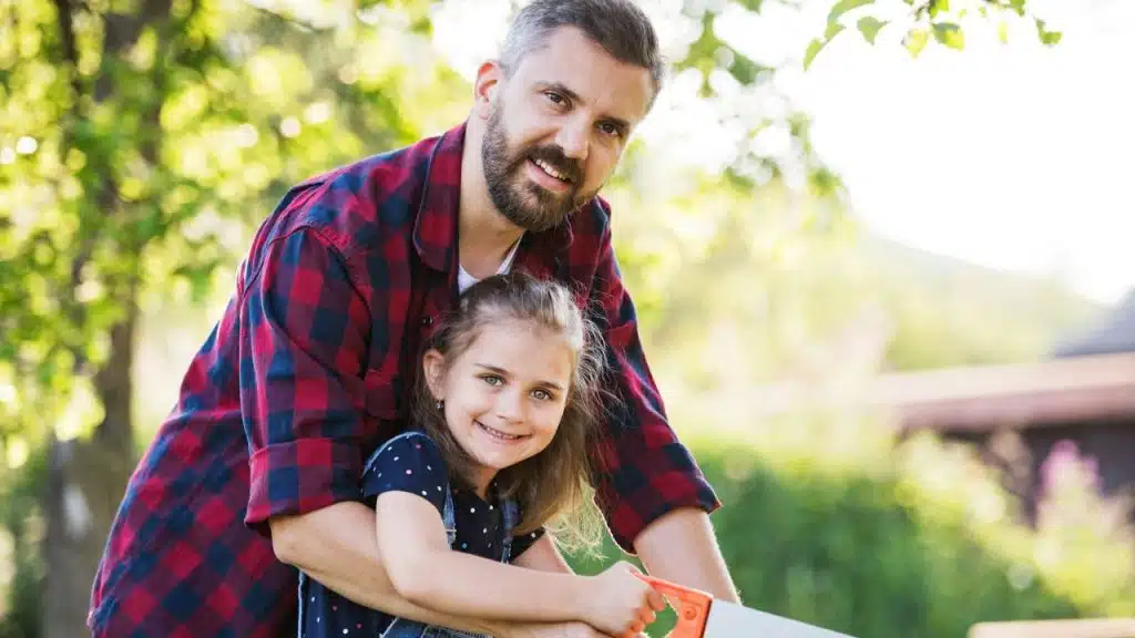 Man in a flannel shirt smiling with a young girl holding a saw outdoors.