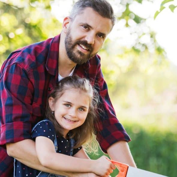 Man in a flannel shirt smiling with a young girl holding a saw outdoors.