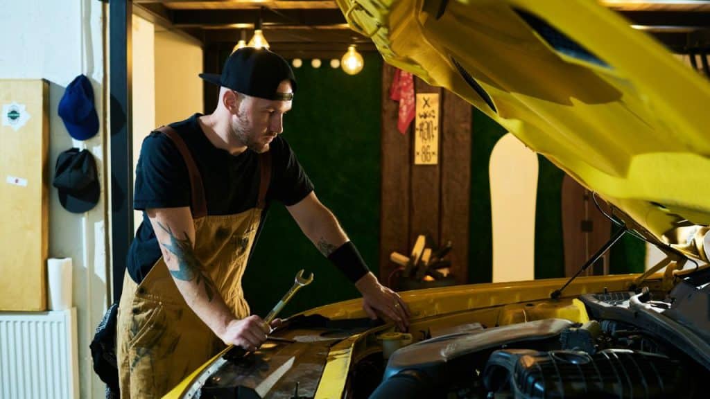 Man in overalls and a cap holding a wrench, working on a yellow car engine.