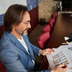 A bespectacled man looking at documents while sitting at a table.