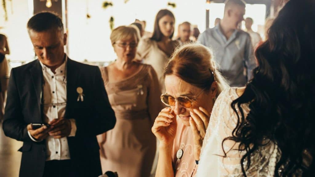 A man in a dark suit looks down at his smartphone while standing in a crowded room with other guests.