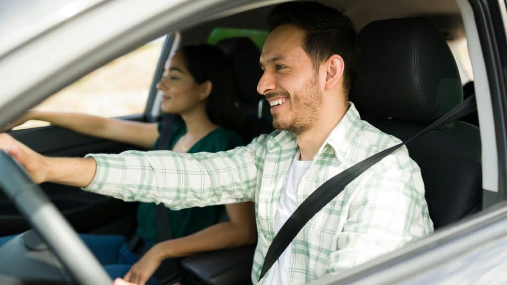 A man wearing a plaid shirt is driving a car and smiling at the passenger sitting next to him.
