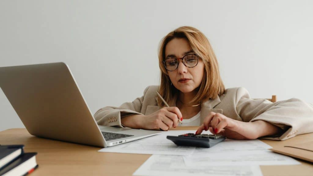 Woman using a calculator and writing notes while looking at a laptop and papers.