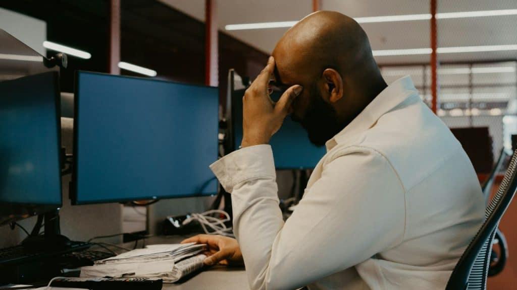 A bald man in a white shirt sits at a desk with two computer screens, holding his head in his hand.